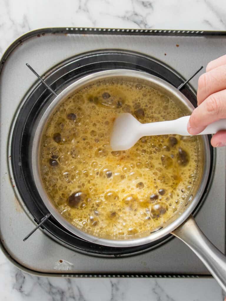 A hand stirs a bubbling golden-brown butter in a saucepan on a stovetop, possibly making caramel or syrup.