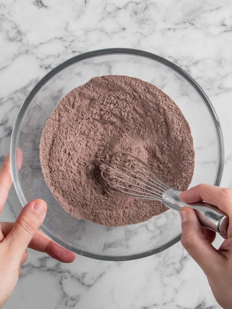 A person whisking a bowl of cocoa powder and dry ingredients on a white marble surface.