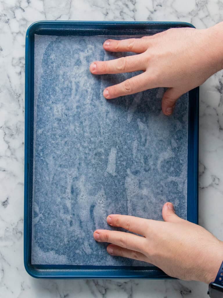 A person’s hands are pressing parchment paper into a blue rectangular baking tray on a white marble surface.