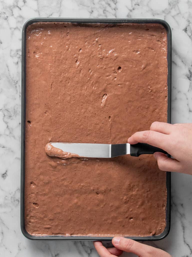 A person uses an offset spatula to spread chocolate cake batter evenly in a rectangular baking pan on a marble surface.