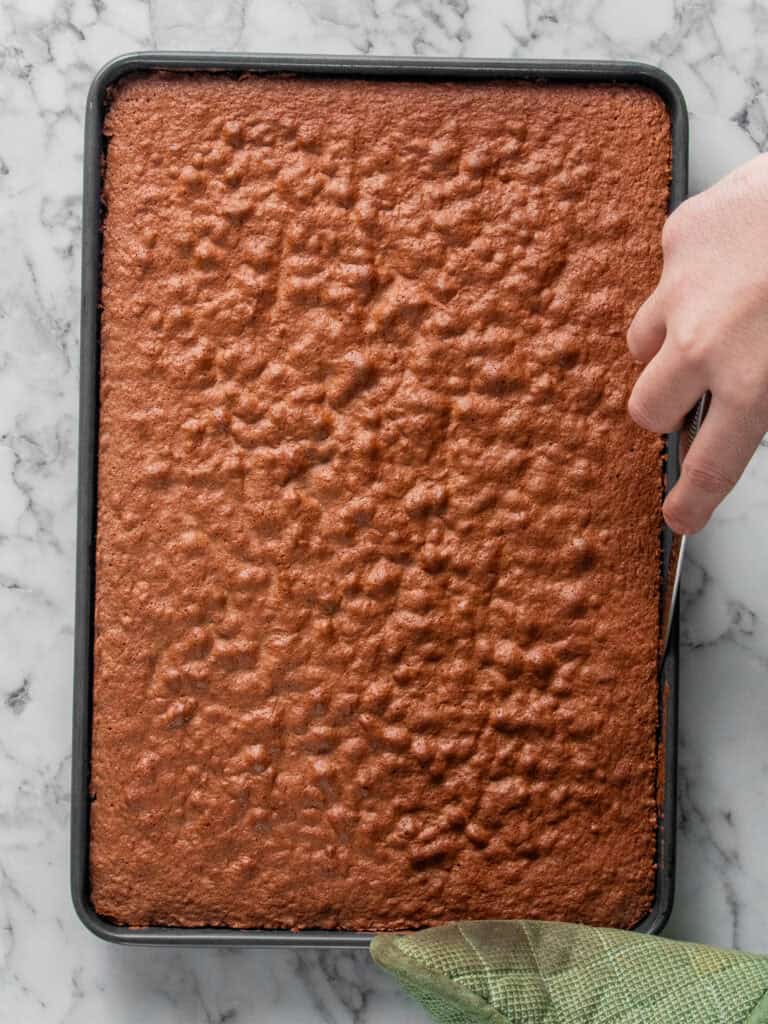 A hand uses a knife to loosen the edge of a large, baked chocolate sheet cake in a rectangular pan on a marble surface, with a green kitchen towel holding the pan.