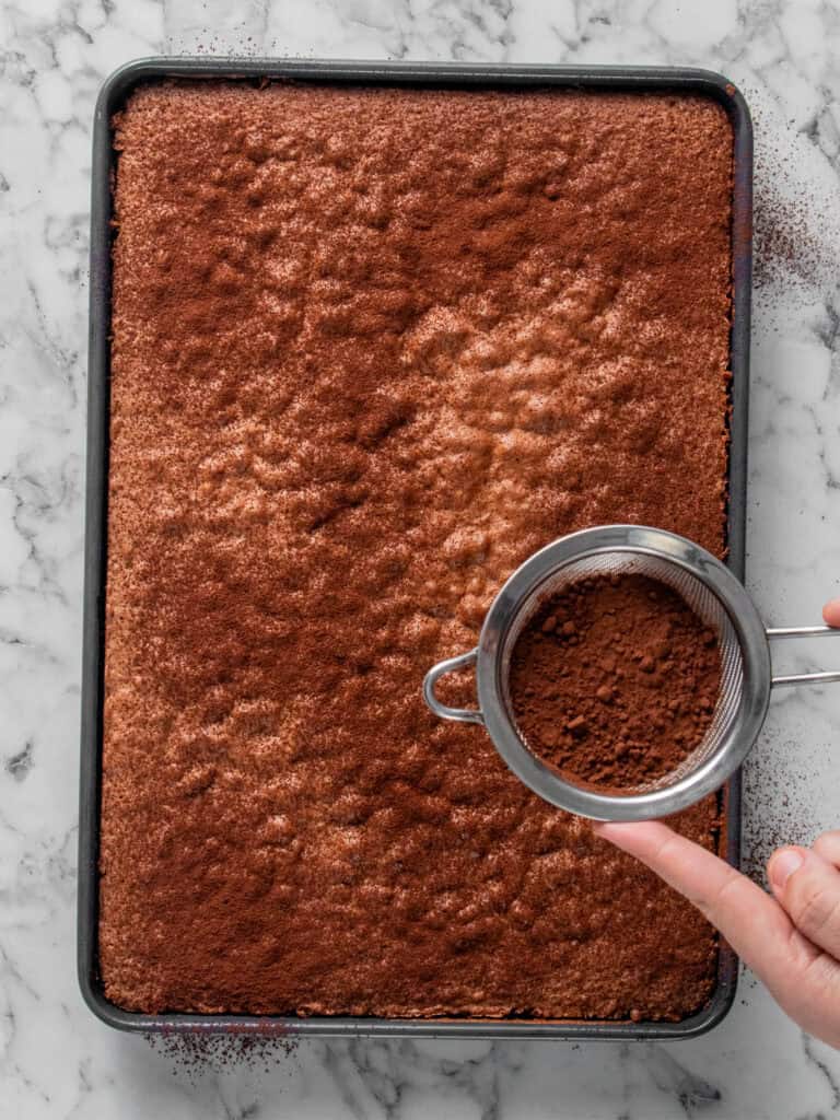 A hand sprinkles cocoa powder through a fine mesh sieve over a rectangular sheet cake on a marble countertop. The cake is covered evenly with the cocoa powder.