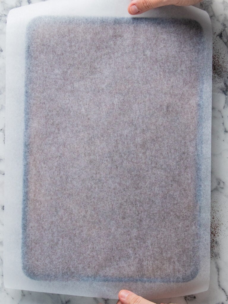 A person holds a baking sheet lined with parchment paper on a marble countertop.