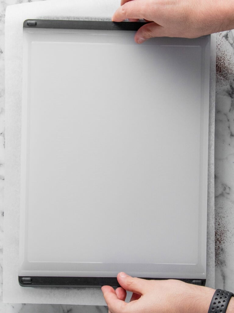 Two hands lift a baking sheet lined with parchment paper on a marble countertop. Only the hands and forearms are visible; the persons left wrist has a dark band or watch.