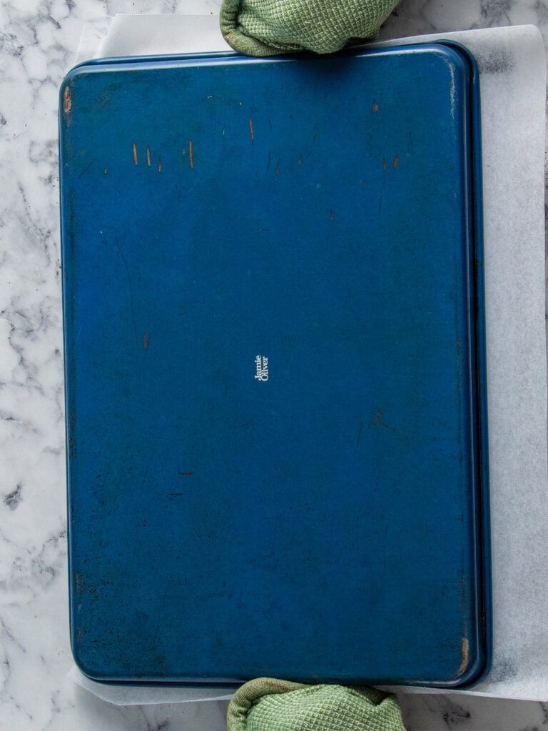 A pair of hands wearing green oven mitts holds a large, blue, rectangular baking sheet upside down on a marble countertop with a sheet of parchment paper underneath.