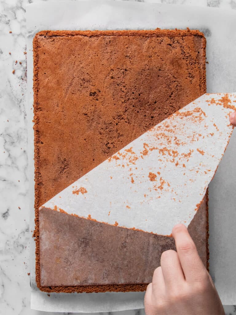 A hand is peeling parchment paper off a large rectangular sheet cake on a marble countertop. The cake is golden brown and the parchment is lined with crumbs.
