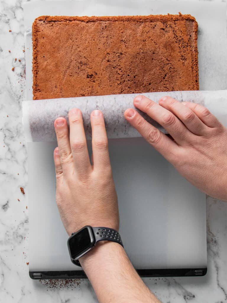 A person rolls up a rectangular sheet cake with parchment paper on a marble surface. The person is wearing a smartwatch on their left wrist.
