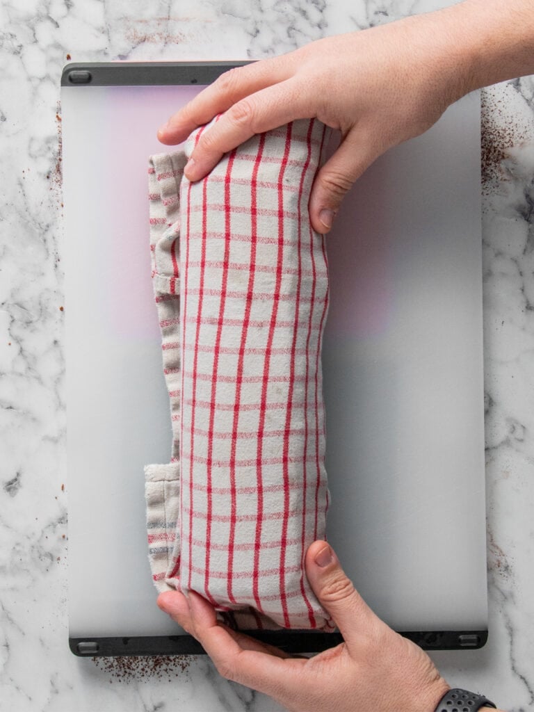 A person wraps a rolled cake in a red and white checkered kitchen towel, placing it on a marble surface with both hands.