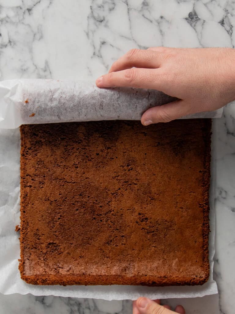 A person’s hands peel parchment paper from a square chocolate cake on a marble countertop.