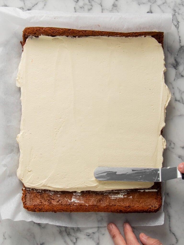 A hand spreads a layer of cream or frosting over a rectangular sheet cake using an offset spatula on a piece of parchment paper, with a marble surface underneath.