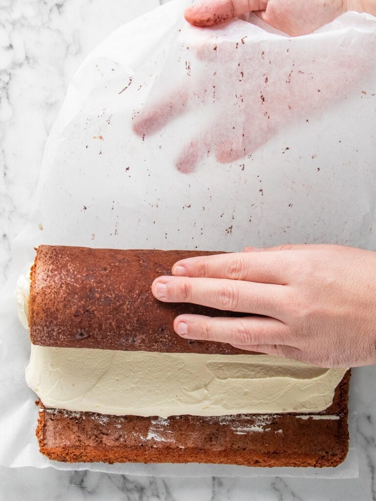 A hand is gently rolling a cake with a layer of white frosting using parchment paper on a marble surface. The cake is being shaped into a roll.