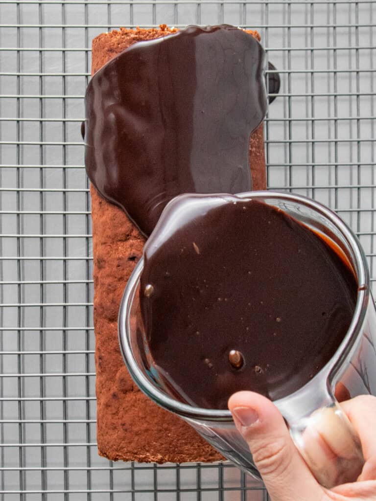 A hand pours glossy chocolate ganache from a glass measuring cup over a chocolate swiss roll cake on a wire cooling rack.