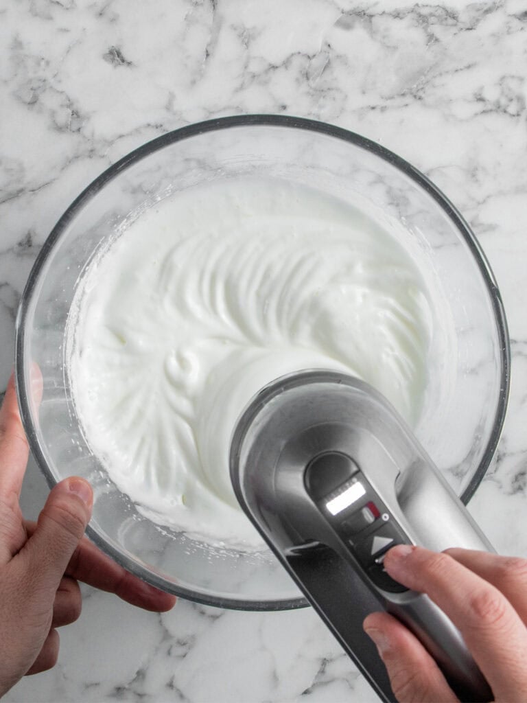 A person uses a hand mixer to whip egg whites in a glass bowl, creating stiff peaks. The bowl is on a white marble surface.