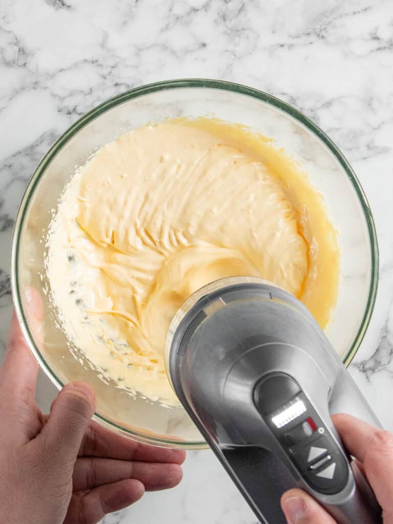 A person uses a handheld electric mixer to blend a creamy, light yellow batter in a glass bowl on a marble countertop.