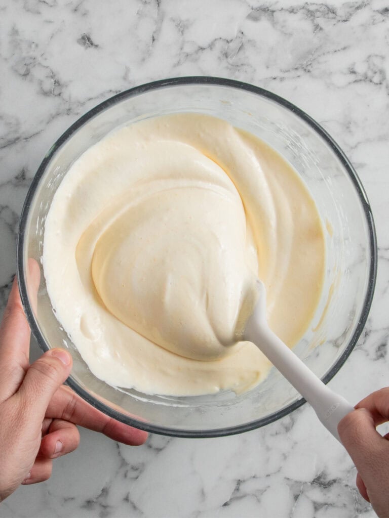 A person stirs a bowl of creamy, pale yellow batter or mixture with a white spatula on a marble countertop.