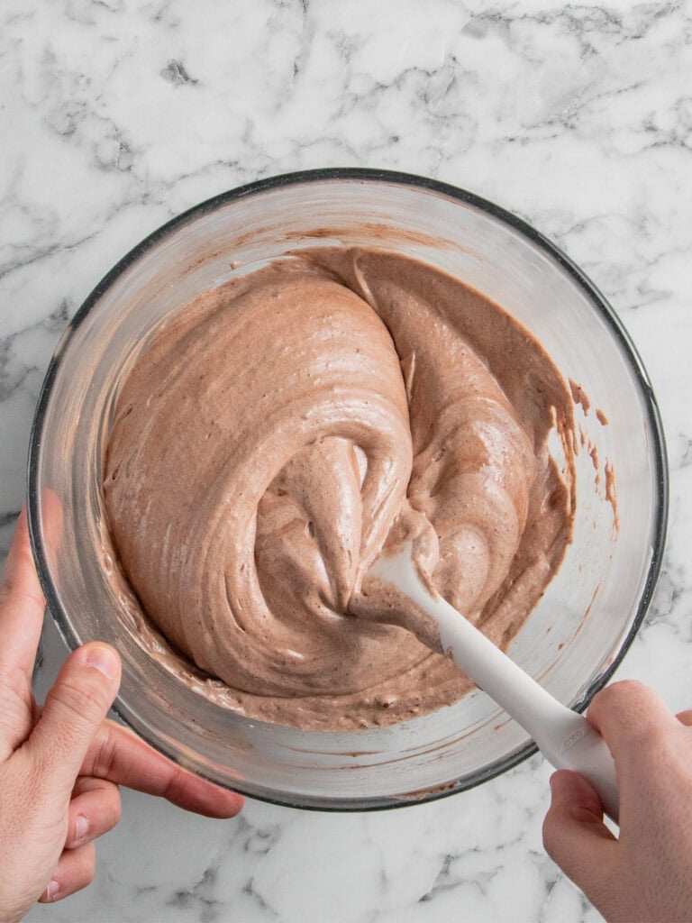 A person mixes light brown cake batter in a glass bowl with a white spatula on a marble countertop.