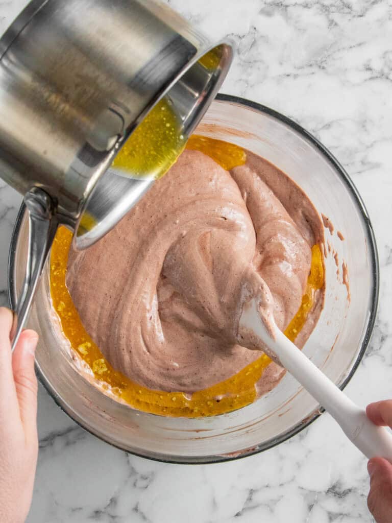 A person pours melted butter from a saucepan into a bowl of chocolate batter, using a spatula to mix, on a marble countertop.