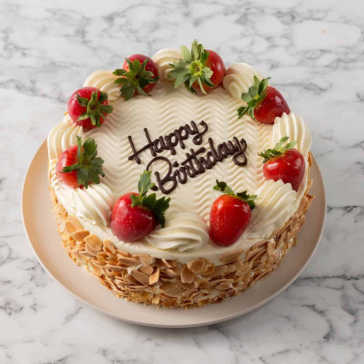 A round cake with white icing, topped with whole strawberries and piped cream, has Happy Birthday written in chocolate. The sides are coated with sliced almonds, and it sits on a white plate on a marble surface.