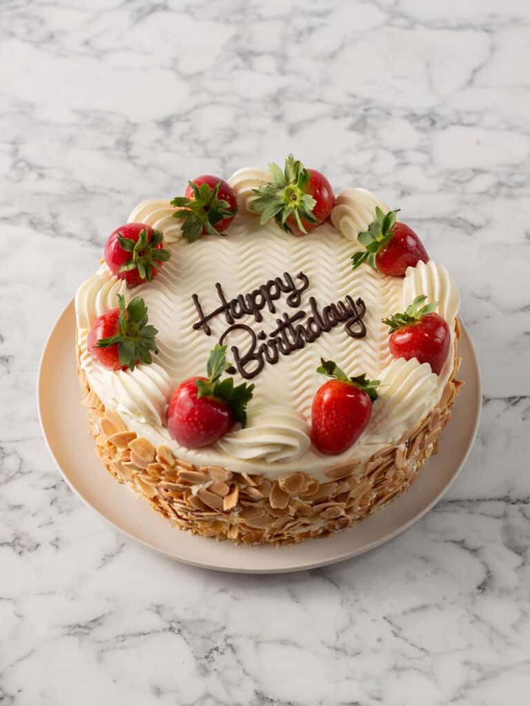 A round birthday cake topped with white frosting, chocolate writing that says Happy Birthday, whole strawberries, and sliced almonds on the sides, sitting on a beige plate on a marble surface.