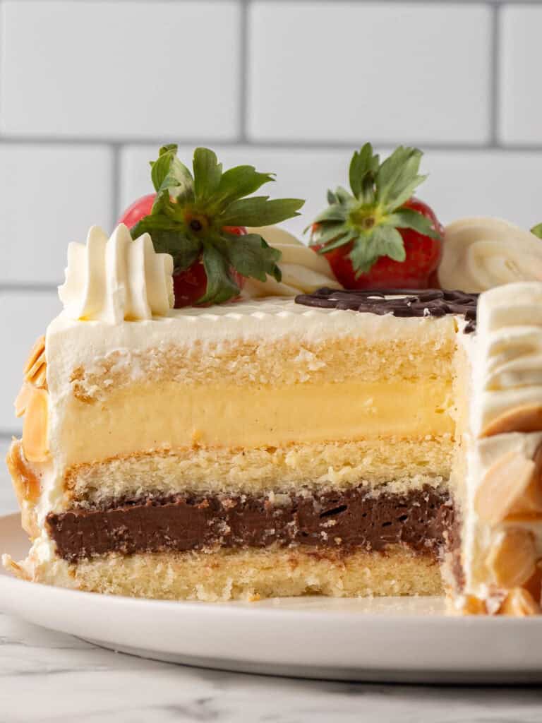 A sliced layer cake with vanilla and chocolate cream layers, topped with white frosting, fresh strawberries, and decorative cream swirls. The cake is on a white plate with a tiled background.