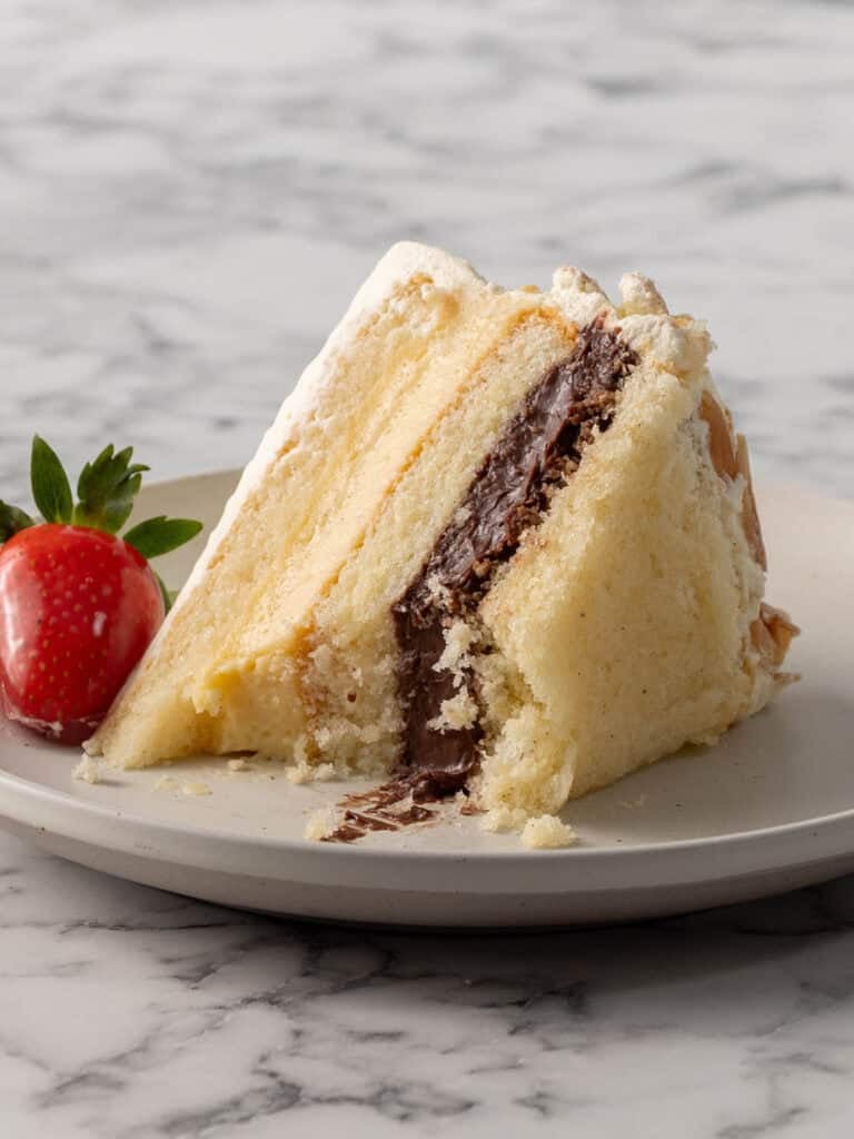 A slice of vanilla layer cake with chocolate filling and white frosting sits on a gray plate, with a fresh strawberry on the side, on a marble surface.
