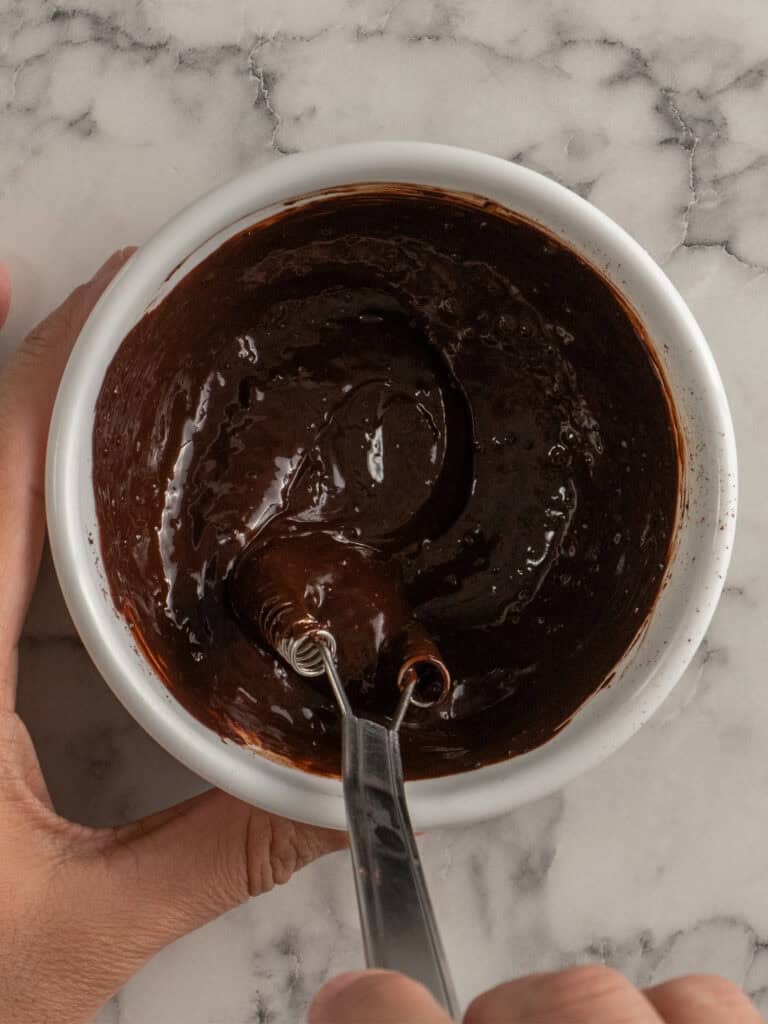 A hand holds a white bowl filled with thick, chocolate and milk being mixed with a small whisk on a marble countertop.