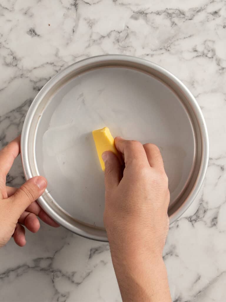 A hand rubs a stick of butter on a round metal cake pan to grease it, on a marble countertop.