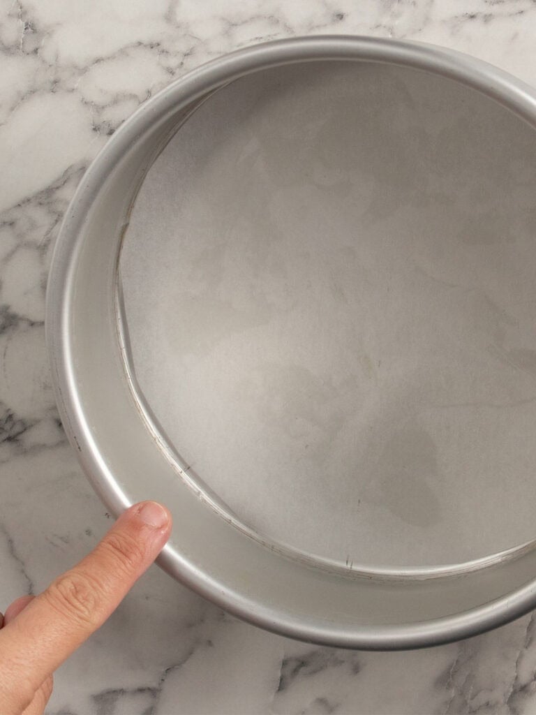 A hand points to a round baking pan lined with parchment paper, set on a marble countertop.