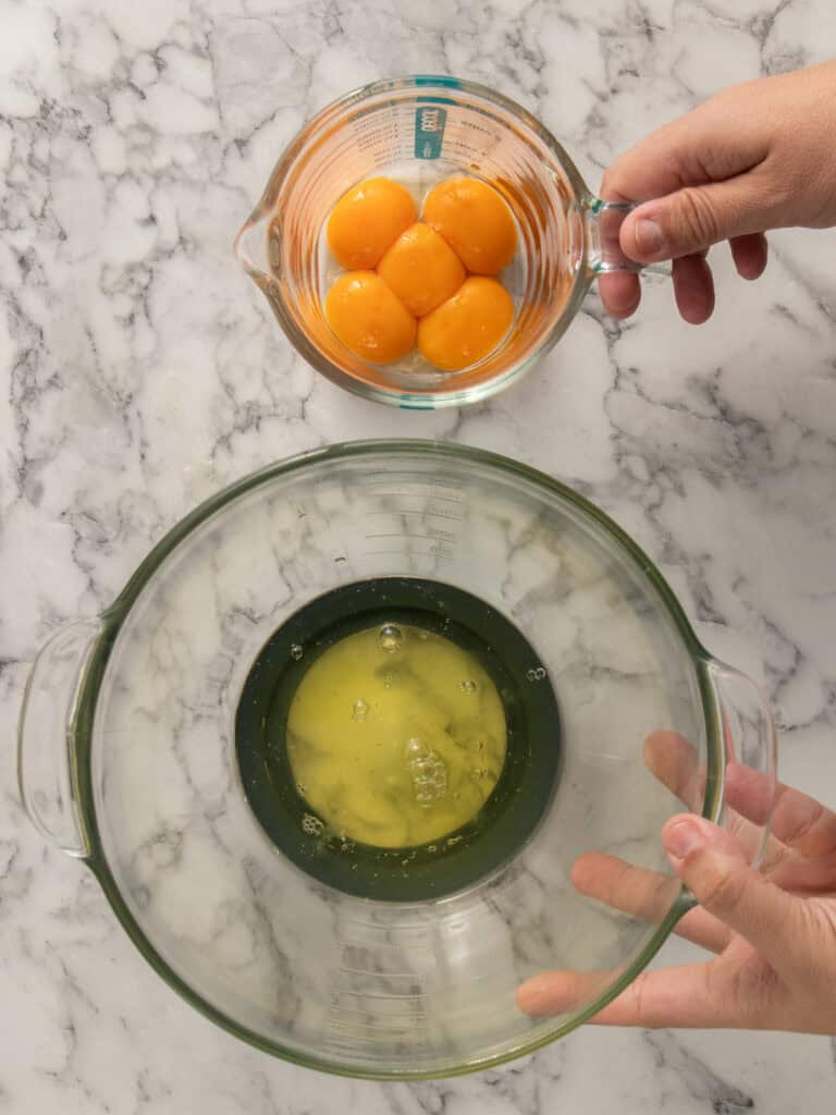 A person holds a small glass measuring cup with egg yolks above a large glass bowl containing egg whites, both on a marble countertop.