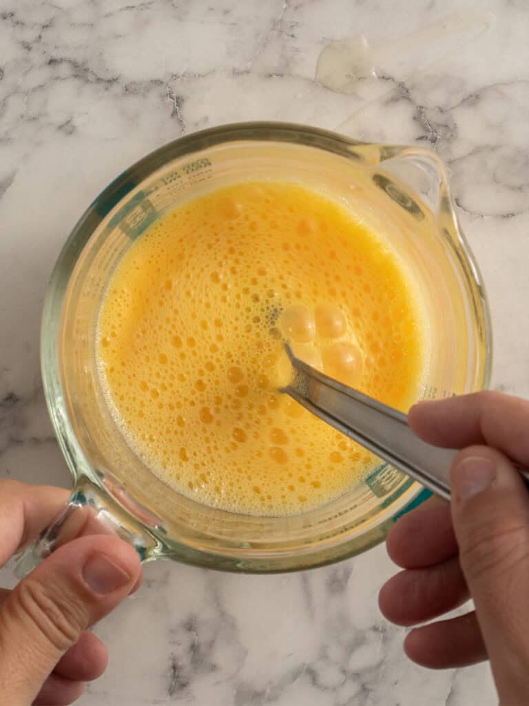 A hand holds a glass measuring cup filled with beaten eggs, mixing them with a metal fork on a white marble countertop.