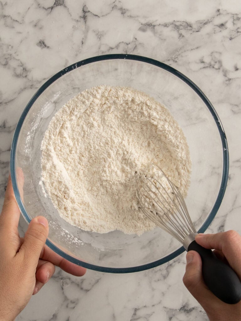 A person whisking flour in a clear glass bowl on a white marble countertop.