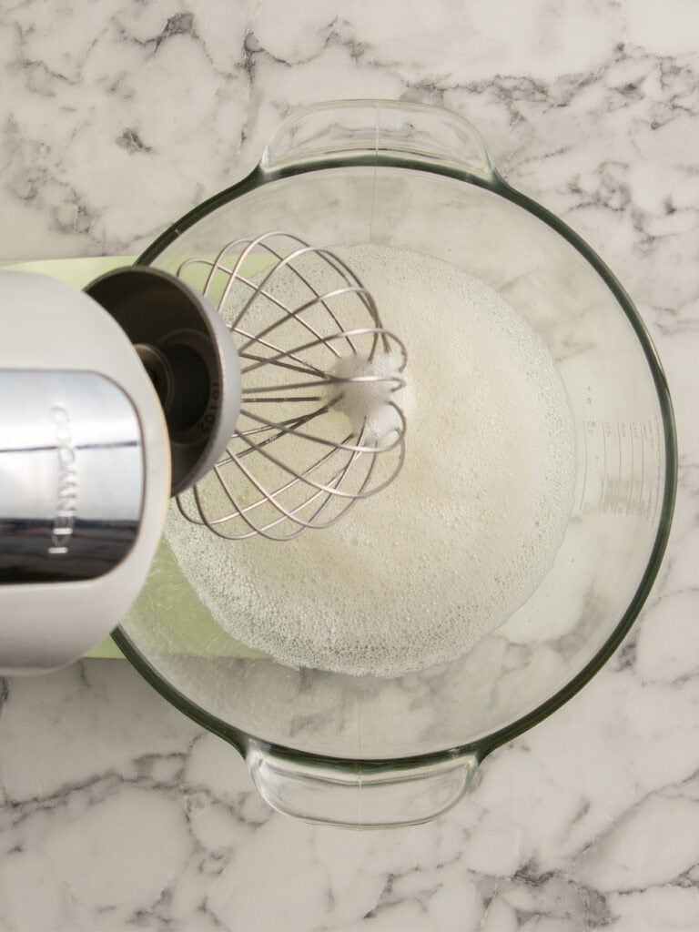 A stand mixer with a whisk attachment is whipping frothy egg whites in a clear glass bowl on a white marble countertop.