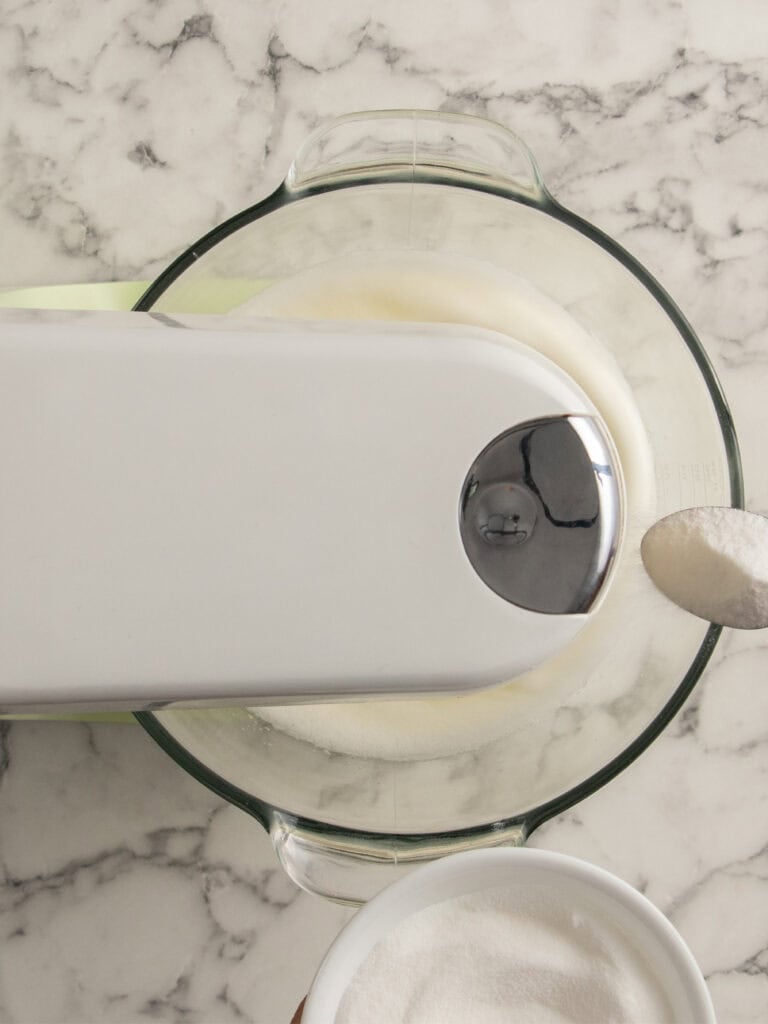 A stand mixer beating a meringue in a glass bowl on a marble countertop, with a spoonful of sugar being added from a white bowl.