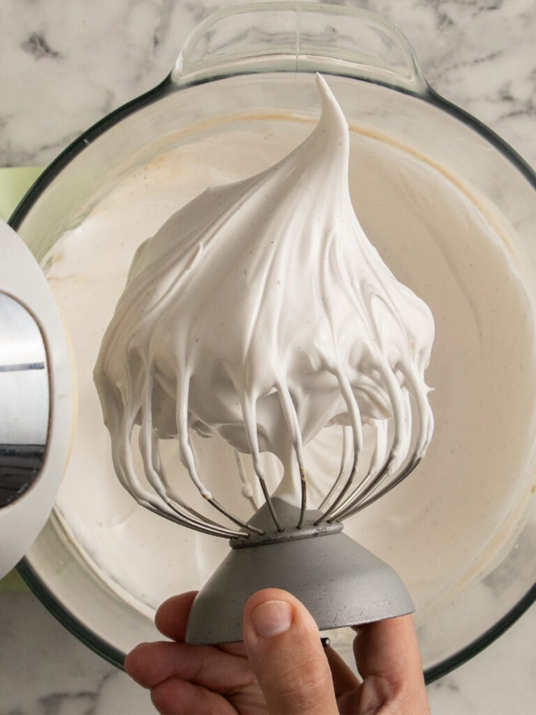A hand holds a stand mixer whisk attachment covered in meringue, stiff peaks of whipped meringue above a glass bowl filled with more meringue, on a marble countertop.