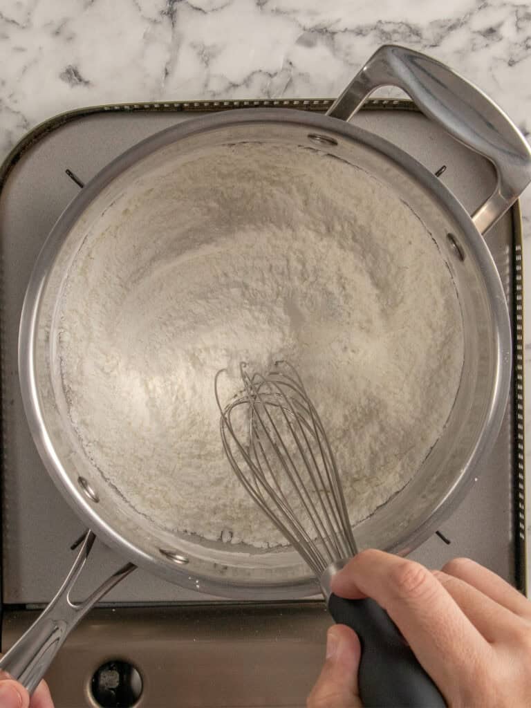 A person whisking white flour in a stainless steel saucepan on a stovetop with a marble countertop in the background.