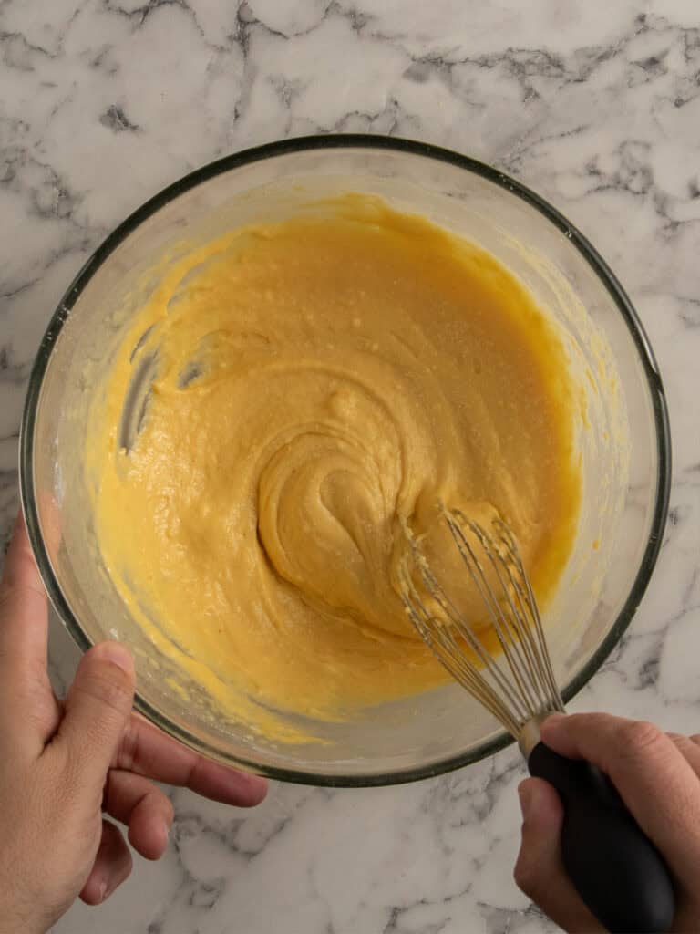 A person whisking yellow cake batter in a glass bowl on a marble countertop.