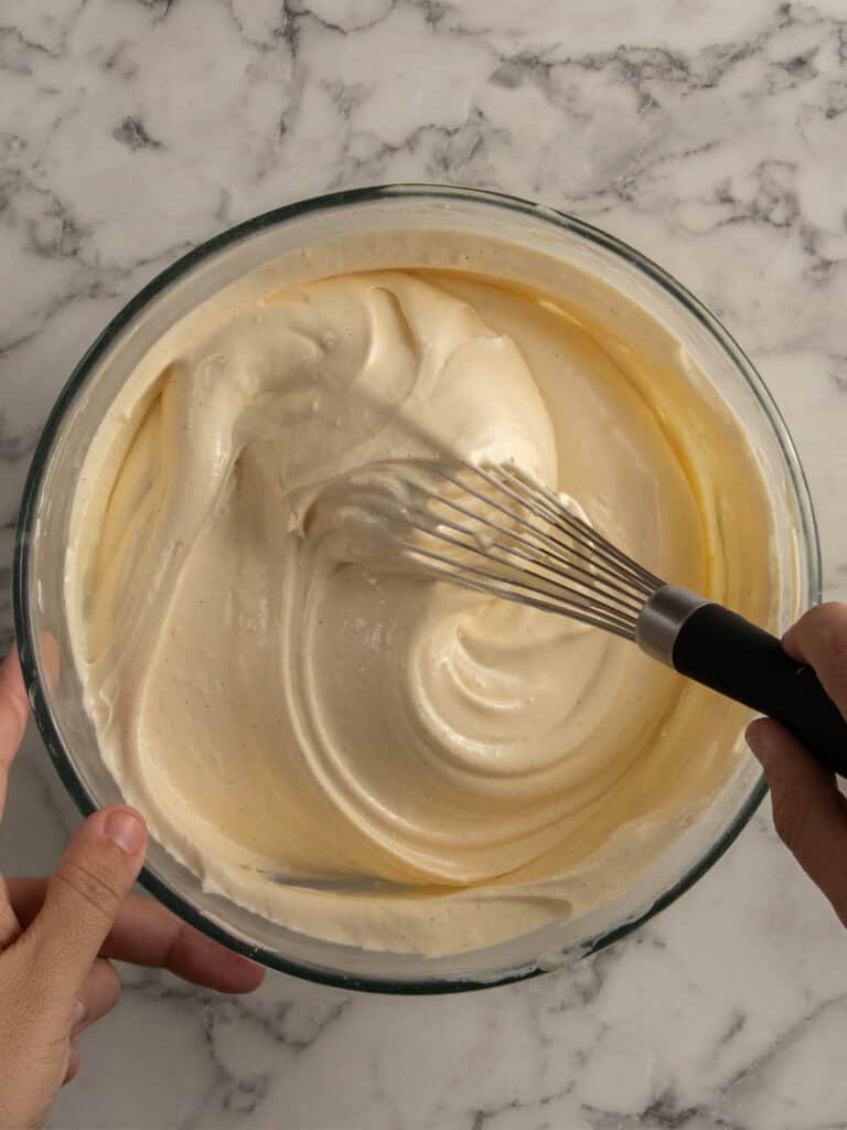A person holds a glass bowl and uses a whisk to mix a creamy cake batter on a marble countertop.