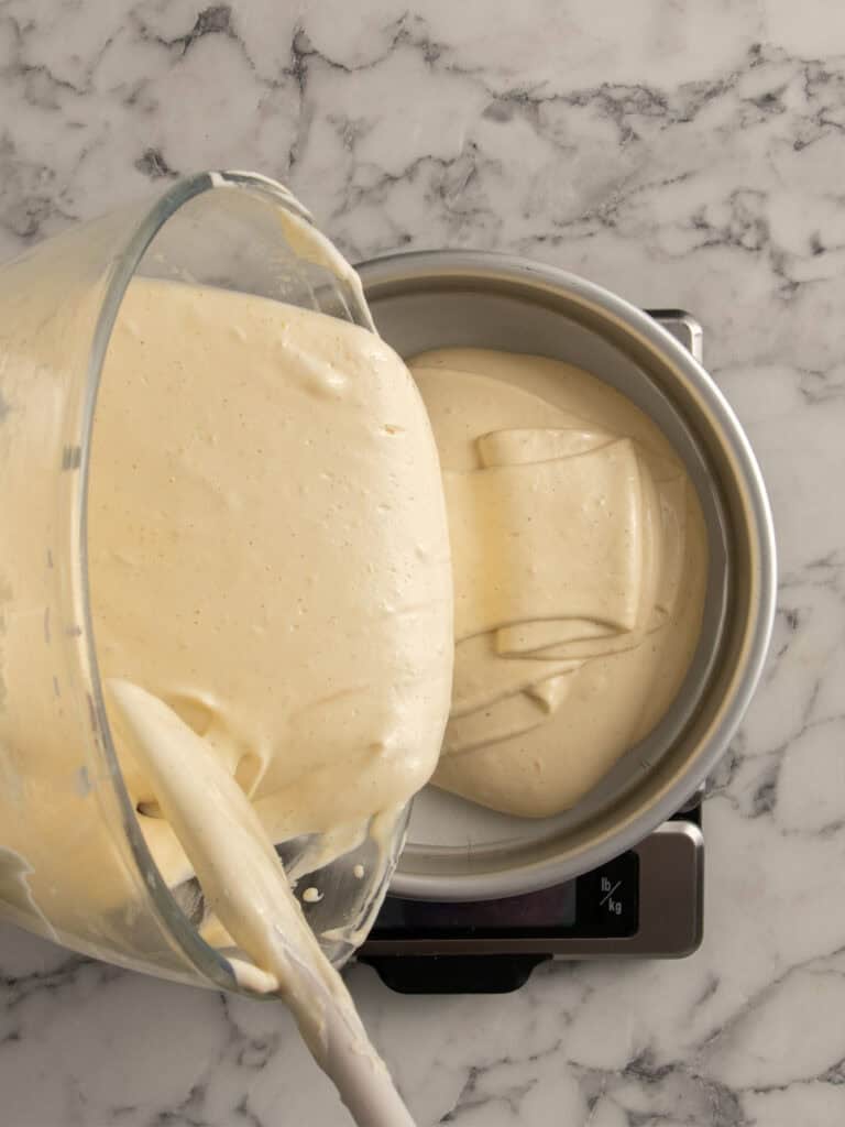 A glass bowl pours fluffy cake batter into a round metal cake pan on a kitchen scale, all set on a white marble countertop.