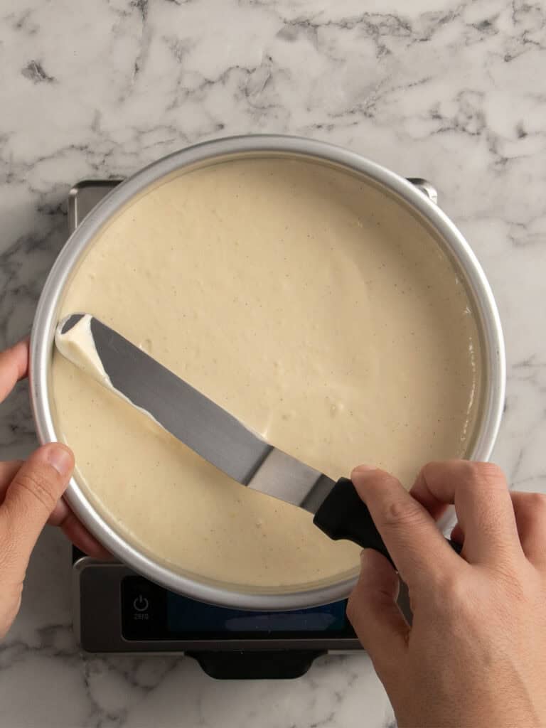 A person uses an offset spatula to smooth cake batter in a round pan on top of a kitchen scale, set on a marble countertop.