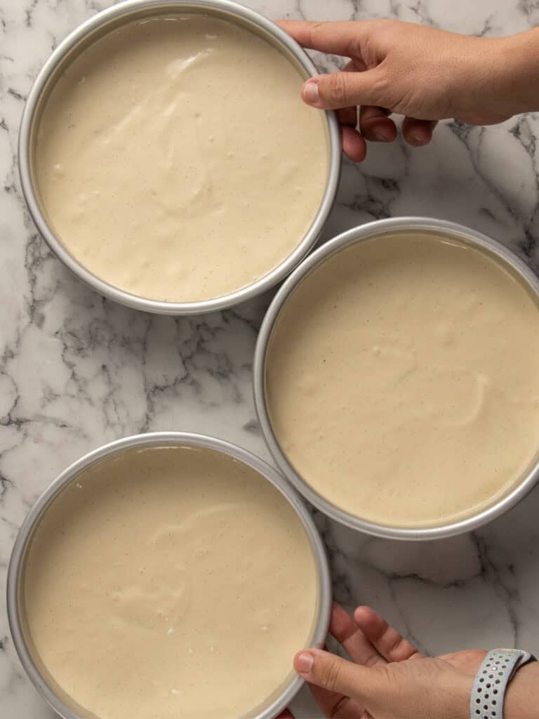 Three round cake pans filled with smooth, pale batter are arranged on a marble countertop, with hands holding two of the pans, preparing them for baking.
