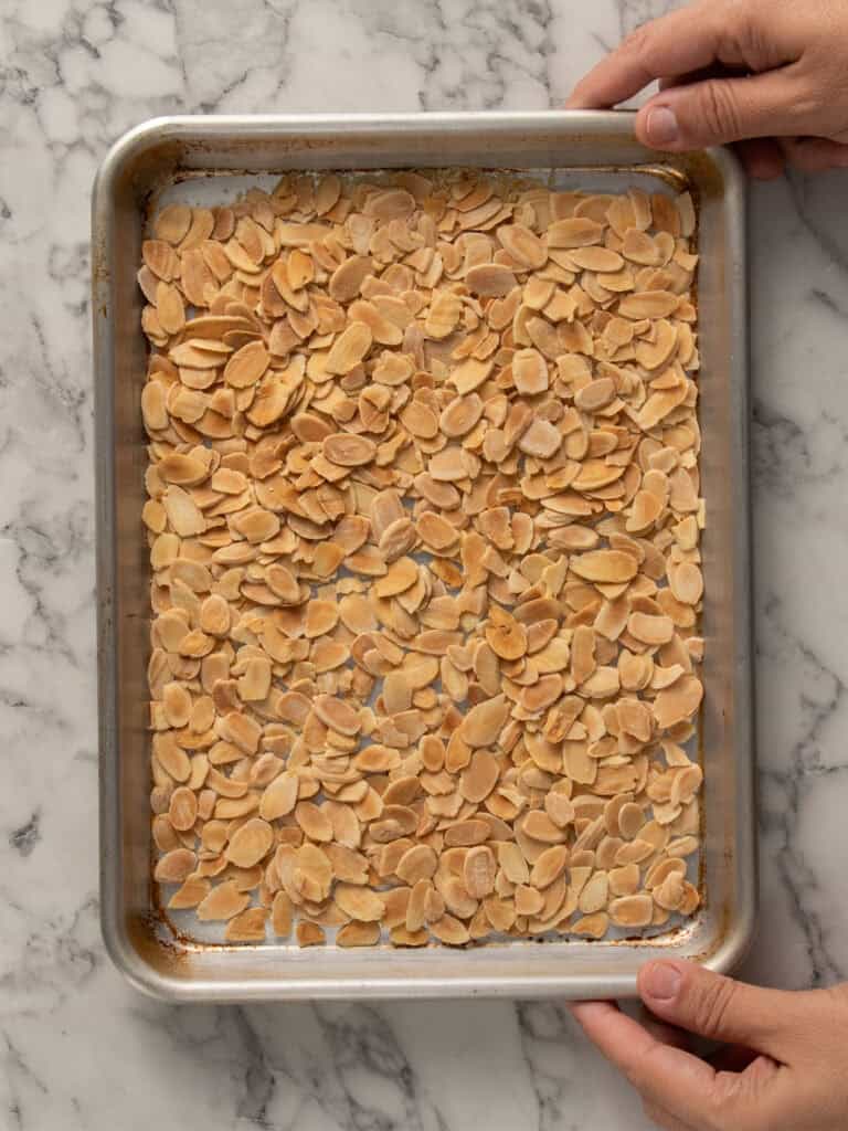 A hand holds a metal baking sheet filled with evenly spread, toasted sliced almonds on a marble countertop.