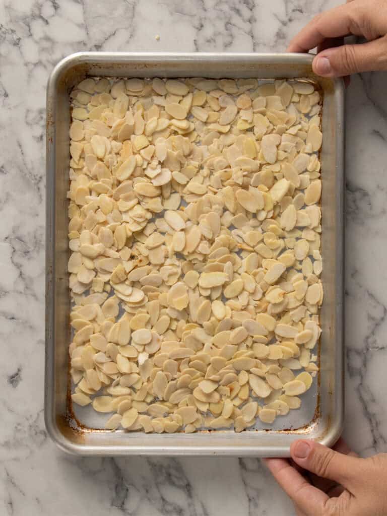 Hands holding a metal baking sheet filled with a single layer of raw sliced almonds, resting on a white marble surface.