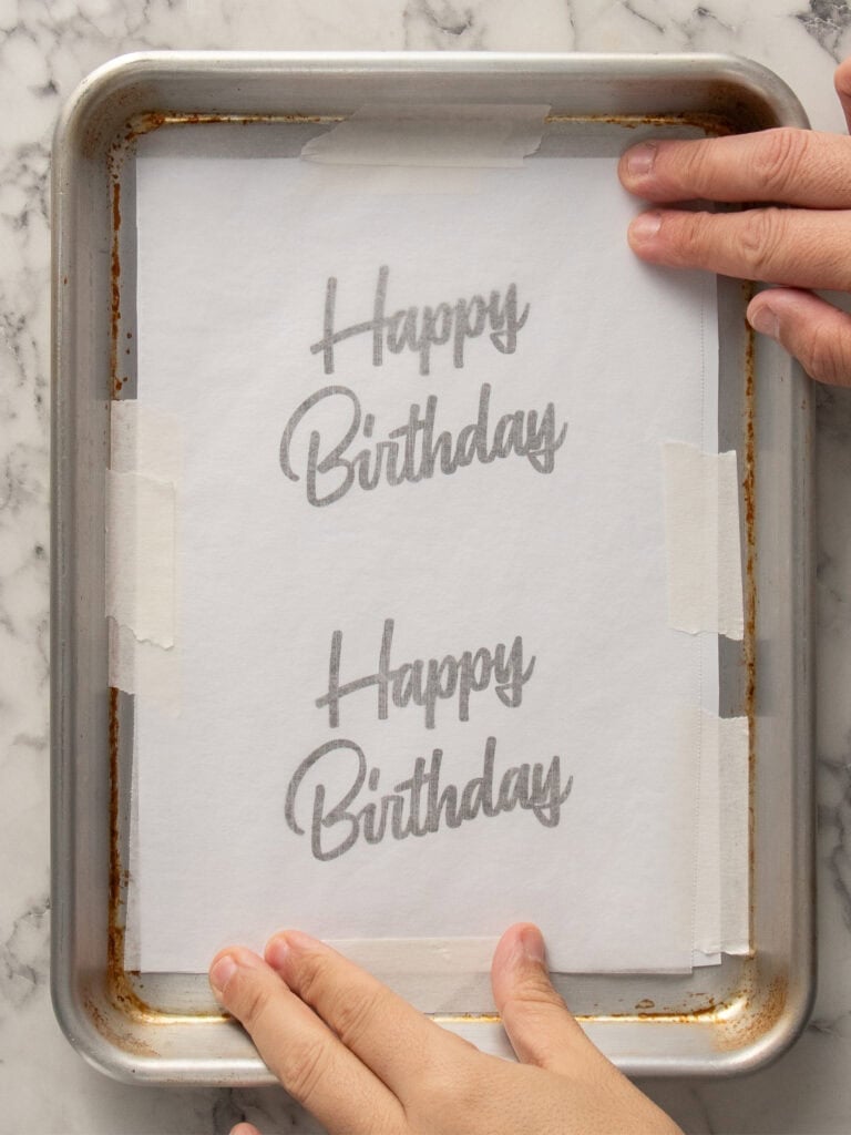A person holds a baking sheet lined with parchment paper, which has Happy Birthday written twice in cursive, with the text visible through the paper. The sheet is placed on a marble surface.