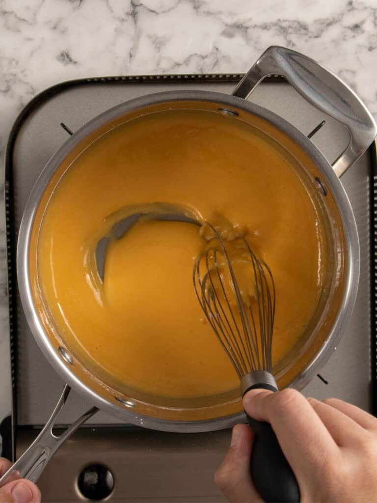 A person whisking egg yolks and milk to make custard in a saucepan on a stovetop with a marble countertop in the background.