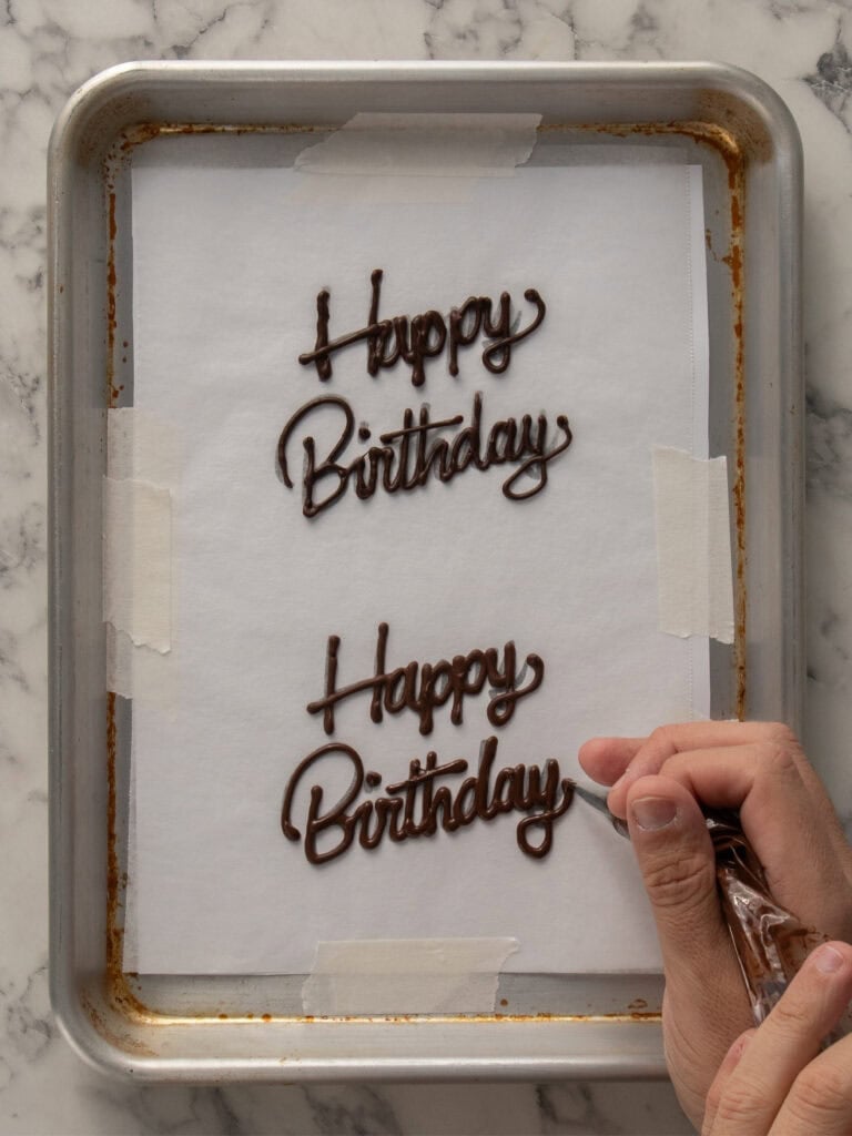 A hand uses a piping bag to write Happy Birthday in chocolate on parchment paper taped to a baking tray. Two chocolate script messages are visible on the tray.