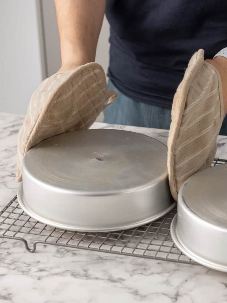 A person wearing oven mitts is inverting a round metal cake pan onto a cooling rack on a marble countertop. Another inverted cake pan is beside it.