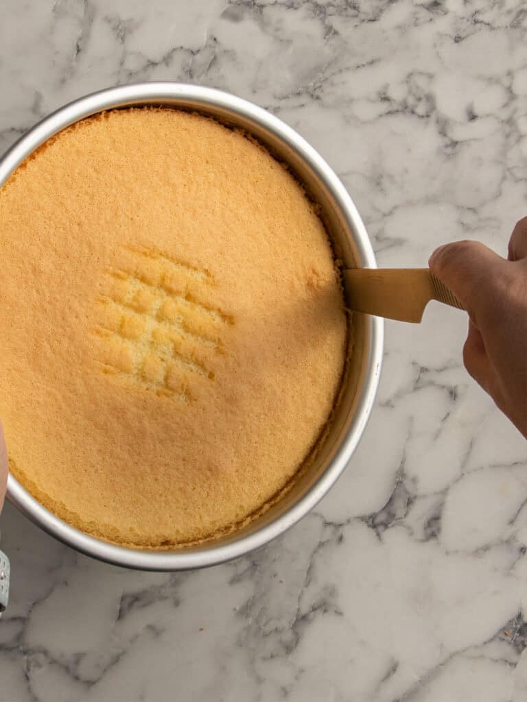 A person uses a small knife to loosen the edge of a round, golden sponge cake in a metal pan on a marble countertop. The cake has a crisscross indentation in the center.