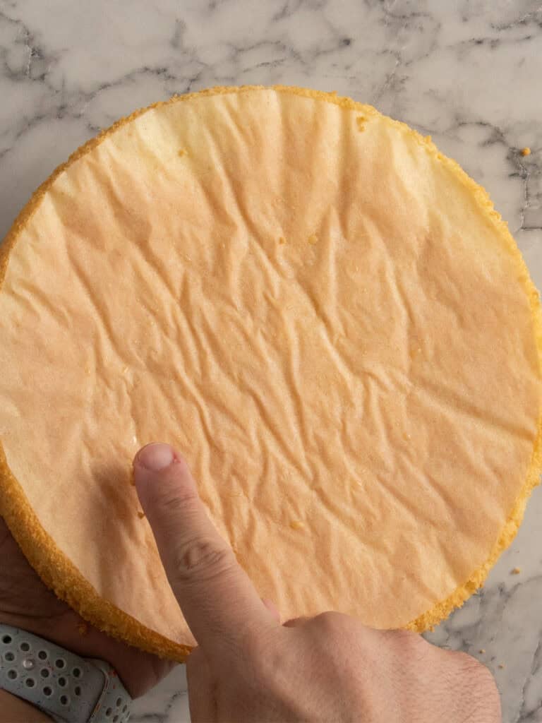 A hand with a smartwatch touches the bottom of a round, golden sponge cake resting on a marble surface.