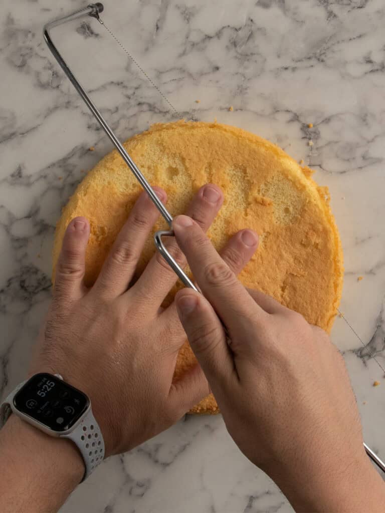 A person uses a wire cake leveler to slice the top off a round yellow cake on a marble surface, steadying the cake with one hand and holding the tool with the other.