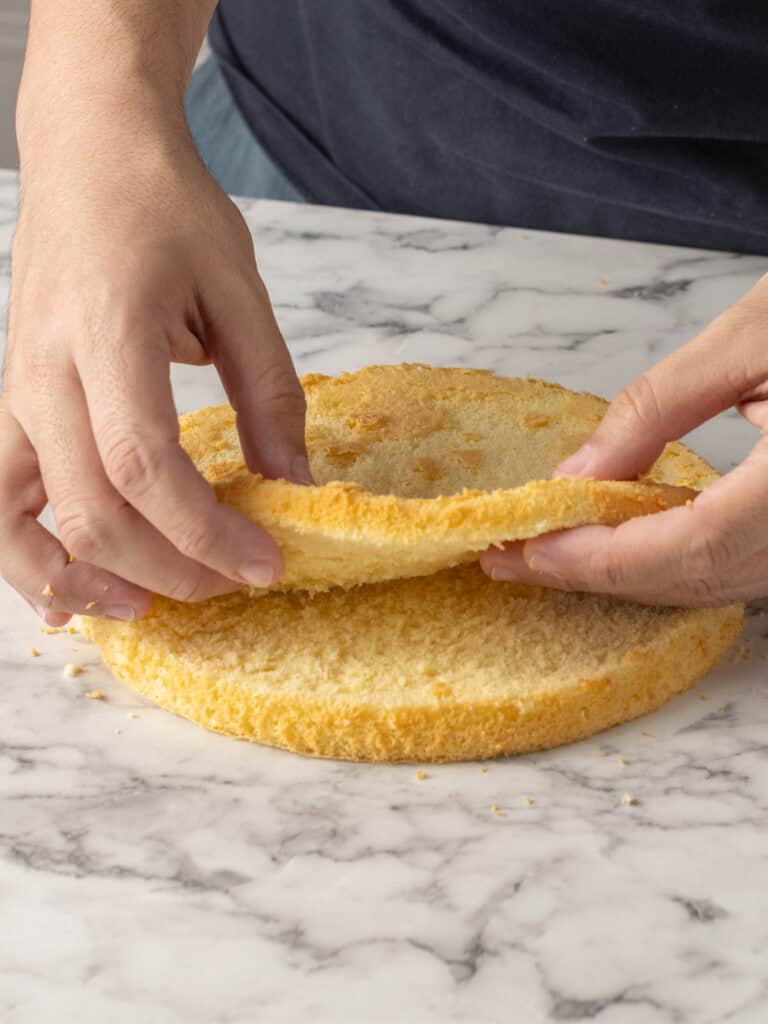 A person uses both hands to separate the top layer from a round sponge cake on a marble countertop.